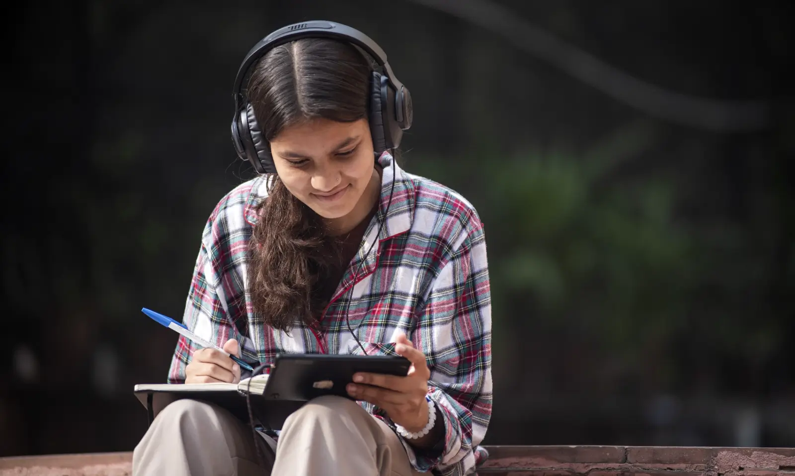 jente med hodetelefoner sitter ute med telefon og tar notater