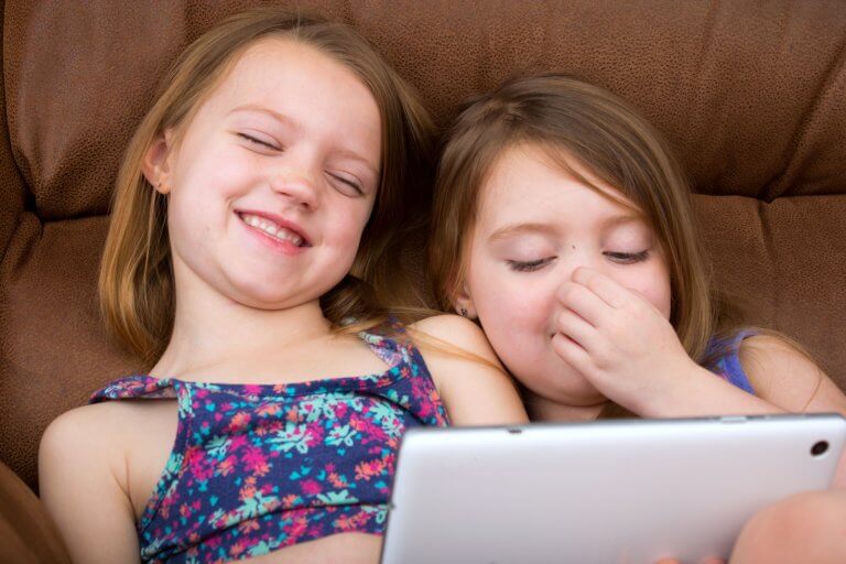 To jenter ligger i en sofaen og ler mens de ser på en ipad